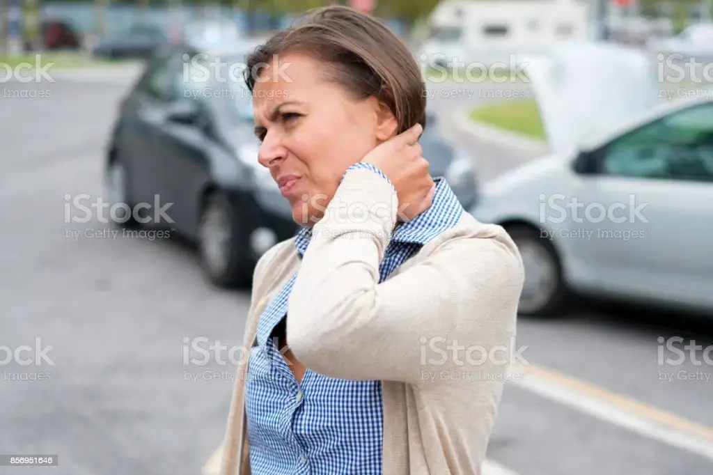 Woman grimacing and holding her neck beside parked cars on a city street